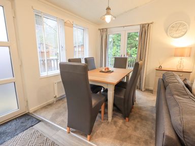 A dining area with a wooden table surrounded by six black chairs next to windows and glass doors with curtains at Cherry Tree Lodge Calgarth 37 White Cross Bay near Windermere