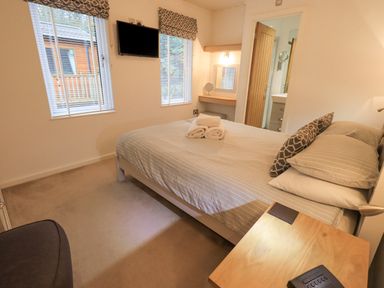 A bedroom with a bed pillows and towels a TV on the wall two windows and a small dressing table at Cherry Tree Lodge Calgarth 37 White Cross Bay near Windermere