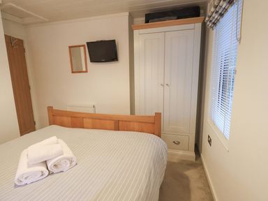 A bedroom with a wooden bed frame with towels on the bed a white wardrobe a wall-mounted television a mirror a wooden door and a window with blinds at Cherry Tree Lodge Calgarth 37 White Cross Bay near Windermere