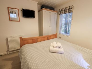 A bedroom with a bed with folded towels on it a wardrobe a window with blinds a radiator a wall-mounted television and a mirror at Cherry Tree Lodge Calgarth 37 White Cross Bay near Windermere