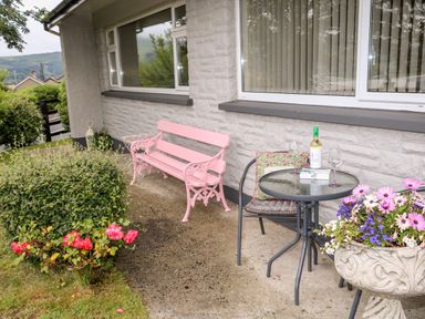 A patio area with a pink bench a round glass table with a bottle of wine and two glasses a chair and flowers in a planter at 27 Pinewood Hill Warrenpoint Northern Ireland