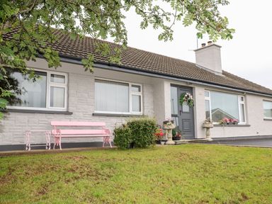 The front exterior of a single-story house with white walls brown roof windows a gray door with a wreath pink bench and flower pots on the lawn at 27 Pinewood Hill in Warrenpoint Northern Ireland