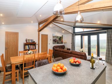 A kitchen island with bowls of fruit and a bottle of wine overlooking a dining area and living room with leather sofas and large windows at Stables Lodge in Wingerworth