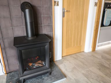 A black wood burning stove with a pipe on a tiled hearth next to a wooden door in a room with light-colored floor tiles at Stables Lodge in Wingerworth