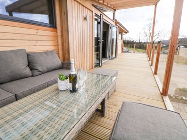 An outdoor wooden deck with a grey cushioned sofa and glass table with a bottle and glasses at Stables Lodge in Wingerworth