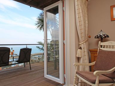 A room with a cushioned wooden chair a side table with a lamp and cream curtains next to a glass door opening to a wooden balcony with chairs and a table overlooking the sea at Wheal Kendall in Mevagissey