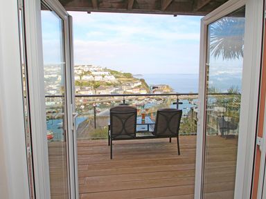 A balcony with two chairs and a table overlooking a harbor and hillside houses at Wheal Kendall in Mevagissey