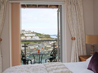 A bedroom with a bed and a bedside table with a lamp looking out to a balcony with a table and two chairs and a view of a harbor at Wheal Kendall in Mevagissey