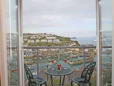 A balcony with a small round table and two chairs overlooking a harbor with boats and houses at Wheal Kendall in Mevagissey