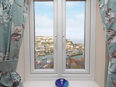 A window with blue floral curtains and a blue bowl on the sill overlooking a harbor with boats and houses at Wheal Kendall in Mevagissey