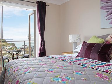 A bedroom with a floral bedspread an open door to a balcony overlooking a coastal town at Wheal Kendall in Mevagissey