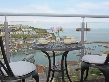 A balcony with a round table and two chairs overlooking a harbor with boats and hillside houses at Wheal Kendall in Mevagissey