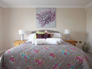 A bedroom with a bed with floral quilt and pillows two bedside tables with lamps and a flower painting on the wall at Wheal Kendall in Mevagissey