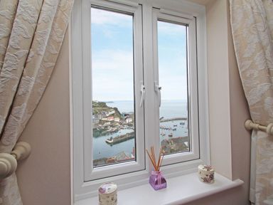A window with curtains and decorative items on the sill overlooking a harbor with boats at Wheal Kendall in Mevagissey