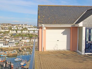 A garage with a closed white door and a blue entrance door next to it overlooking a harbor with boats and houses on a hillside at Wheal Kendall in Mevagissey