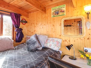 A wooden bedroom with a bed covered in pillows and a blanket next to a window with purple curtains a small table with a lamp and flowers and a wall mirror at Packhorse Shepherd's Hut in Alderwasley near Wirksworth