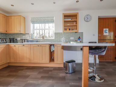 A kitchen with wooden cabinets a window above the sink a wall clock a wooden door and a counter with bar stools at Ilgram in St Newlyn East