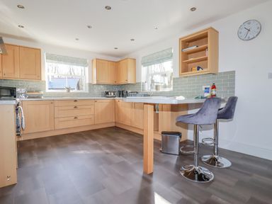 A kitchen with wooden cabinets gray tiled backsplash two windows and two gray bar stools at Ilgram in St Newlyn East