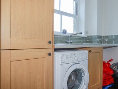 A laundry room with a front-loading washing machine under a countertop with a sink and wooden cabinets at Ilgram in St Newlyn East