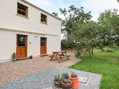 An outdoor area with two wooden doors on a white building a wooden picnic table gravel and grass with trees and several potted plants near Bramblewood Cottage in Crookdake near Bassenthwaite