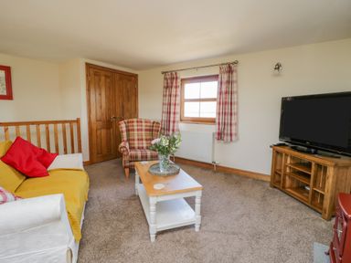 A living room with a sofa and armchair around a coffee table with flowers a television on a wooden stand and a window with plaid curtains at Bramblewood Cottage in Crookdake near Bassenthwaite