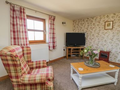 A living room with a plaid armchair a wooden coffee table with flowers a television on a stand and floral wallpaper at Bramblewood Cottage in Crookdake near Bassenthwaite