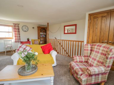 A sitting area with a plaid armchair and sofa with yellow cover a wooden coffee table with flowers and a bookshelf at Bramblewood Cottage in Crookdake near Bassenthwaite