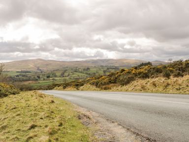 A countryside road with grassy banks and hills in the background at Bramblewood Cottage in Crookdake near Bassenthwaite