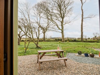 A wooden picnic table with a bottle and two glasses on a gravel patio with trees and grass beyond at Bramblewood Cottage in Crookdake near Bassenthwaite