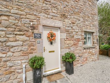 A stone exterior with a cream door decorated with a floral wreath and two potted plants at Bella Cottage Bolton near Appleby-in-Westmorland