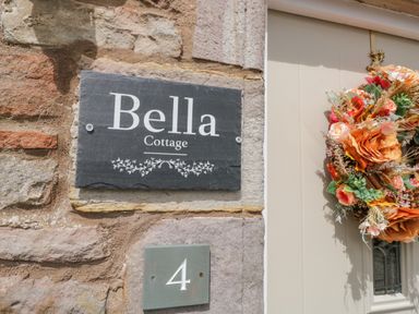 A stone wall with a plaque that reads Bella Cottage a green square plaque with number 4 and a beige door with a floral wreath at Bella Cottage in Bolton near Appleby-in-Westmorland