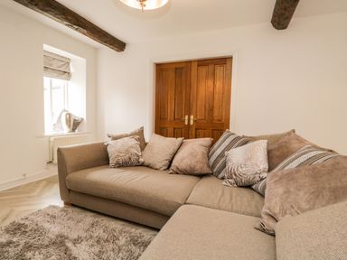 A living room with a beige sectional sofa with patterned cushions a wooden double door and a window with blinds at Bella Cottage Bolton near Appleby-in-Westmorland