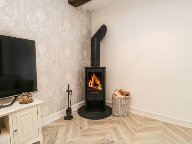 A living room corner with a black wood-burning stove a basket of firewood a white cabinet with a television and patterned wallpaper at Bella Cottage Bolton near Appleby-in-Westmorland