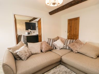 A living room with a beige L-shaped sofa and multiple cushions next to a wooden door and an open doorway to a kitchen at Bella Cottage Bolton near Appleby-in-Westmorland