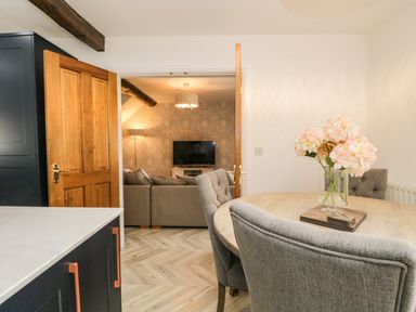 A dining area with a round wooden table and grey upholstered chairs with flowers on the table and a view into a living room with a sofa and a television at Bella Cottage Bolton near Appleby-in-Westmorland