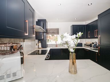 A kitchen with dark cabinets a white marble countertop a vase with white flowers a window above the sink and various kitchen appliances at Bella Cottage in Bolton near Appleby-in-Westmorland