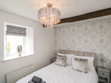 A bedroom with a bed two nightstands a chandelier patterned wallpaper and a window with a vase on the sill at Bella Cottage in Bolton near Appleby-in-Westmorland
