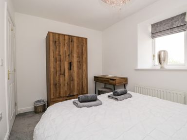 A bedroom with a bed, wooden wardrobe, desk, window with vase, and rolled towels on the bed at Bella Cottage Bolton near Appleby-in-Westmorland
