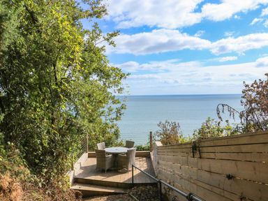 A wooden deck with a round table and four chairs overlooking the sea surrounded by trees and a wooden fence at Sail Loft in Dawlish
