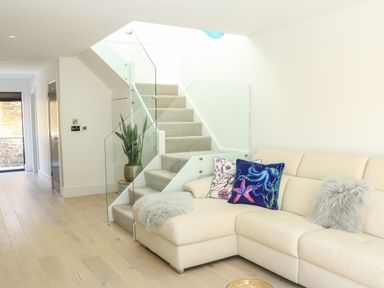 A living area with a beige sectional sofa decorated with cushions next to carpeted stairs with glass railing and a potted plant at Sail Loft in Dawlish