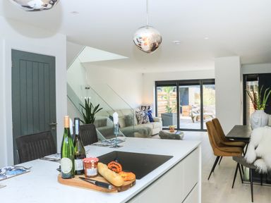 A kitchen island with bottles and bread near a dining table and living area with sofa and glass doors at Sail Loft in Dawlish