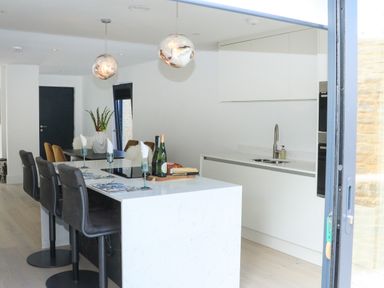 A kitchen with a white island and dark chairs alongside a dining table with yellow chairs and plants at Sail Loft in Dawlish
