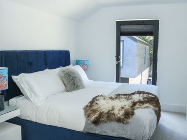 A bedroom with a blue upholstered bed a white comforter fluffy pillows a window and a nightstand with a lamp at Sail Loft in Dawlish