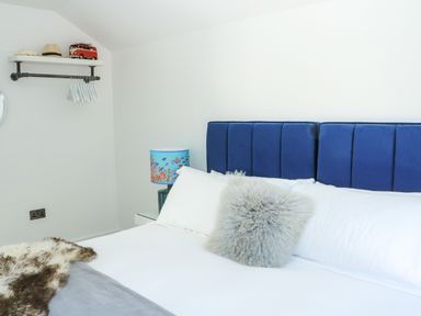 A bedroom with a blue headboard white bed linens a gray pillow a lamp with fish design and a shelf with hats and clothing hangers at Sail Loft in Dawlish
