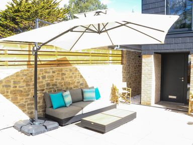 An outdoor patio with a grey sofa and blue cushions under a large umbrella near a black door at Sail Loft in Dawlish