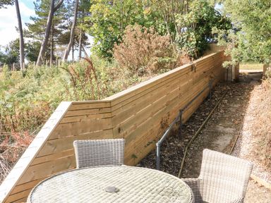 A round glass top table with wicker chairs on a patio next to a wooden fence and greenery at Sail Loft in Dawlish