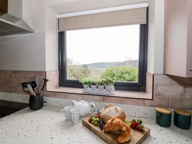 A kitchen countertop with a teapot, two mugs, bread, croissant, fruit, utensil holder, and plants on the windowsill at Sevenwoods View in Longhope