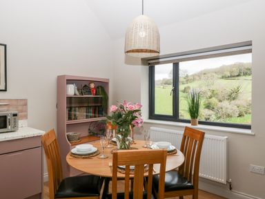A dining area with a round wooden table set with plates glasses and pink flowers near a window showing a green landscape at Sevenwoods View in Longhope