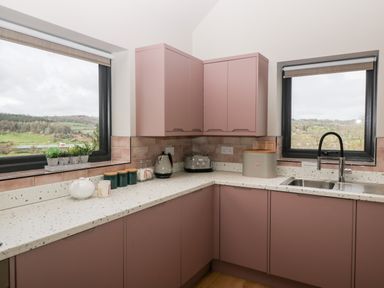 A kitchen with mauve cabinets a sink toaster kettle containers and two windows showing countryside at Sevenwoods View in Longhope