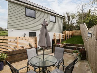 An outdoor patio area with a glass table and four chairs a closed umbrella and potted plants beside wooden fencing and a grassy raised garden at Sevenwoods View in Longhope